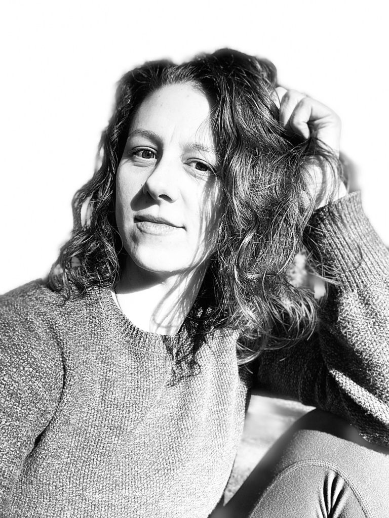 A black and white headshot of Kayla with wavy hair, wearing a textured sweater, sitting in natural light against a bright background.