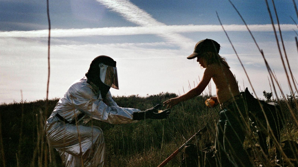 A person in a reflective suit kneels in a grassy field, reaching toward a child under a sky marked with cloud trails, suggesting environmental observation and interaction. The scene represents an exploration of cutting-edge weather modification technology designed to enhance public safety and environmental management through effective atmospheric intervention.