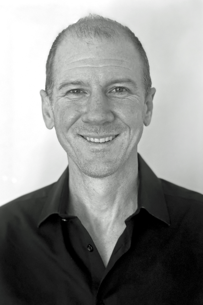 A black and white headshot of Sean smiling, wearing a dark collared shirt against a neutral background.